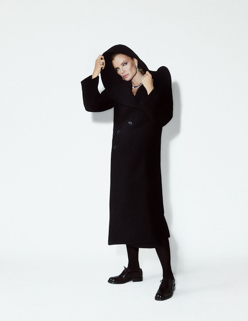 Full-length portrait of a stylish adult woman in a hooded black coat, black tights, and black shoes against a stark white background.