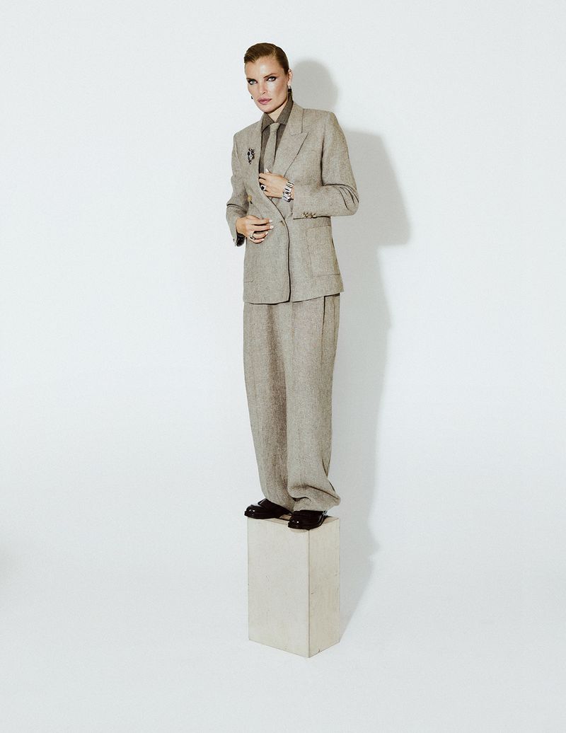 Adult woman stands on white block in light brown suit with dark shirt and tie in photo studio against white backdrop with neutral lighting.
