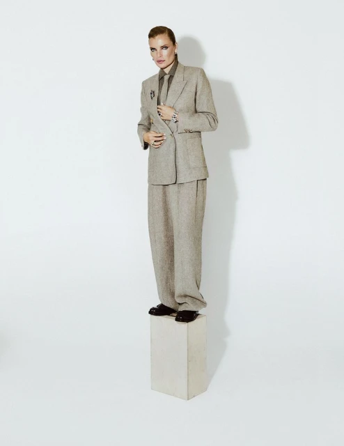 Adult woman stands on white block in light brown suit with dark shirt and tie in photo studio against white backdrop with neutral lighting.
