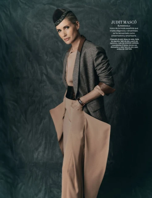 Elegant woman in a tan pantsuit and patterned blazer, accented by a black belt and decorative hat, poses against a muted gray backdrop.