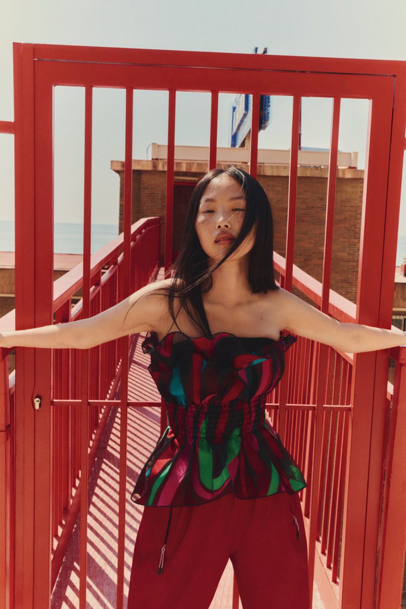 A stylish Asian woman in a colorful strapless top and red pants poses on a red staircase, against a coastal building and ocean backdrop.