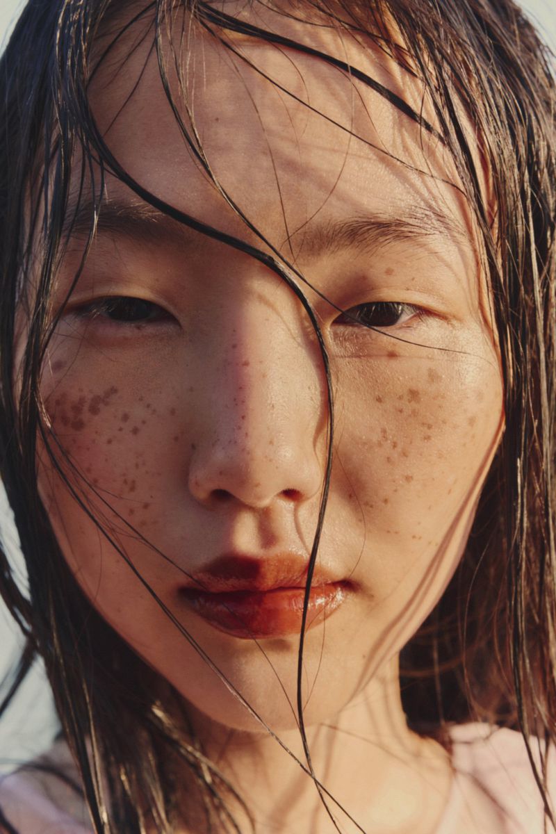 Close-up portrait of a young adult woman with freckles and wet hair, face illuminated by natural light, glossy lips, and fair skin.