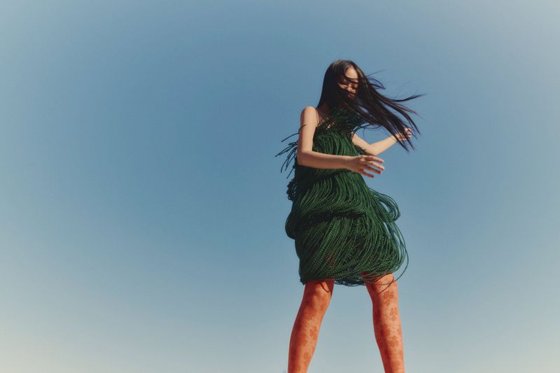 Woman in green fringe dress with patterned tights poses against a clear blue sky in outdoor fashion shot, wind blowing hair.