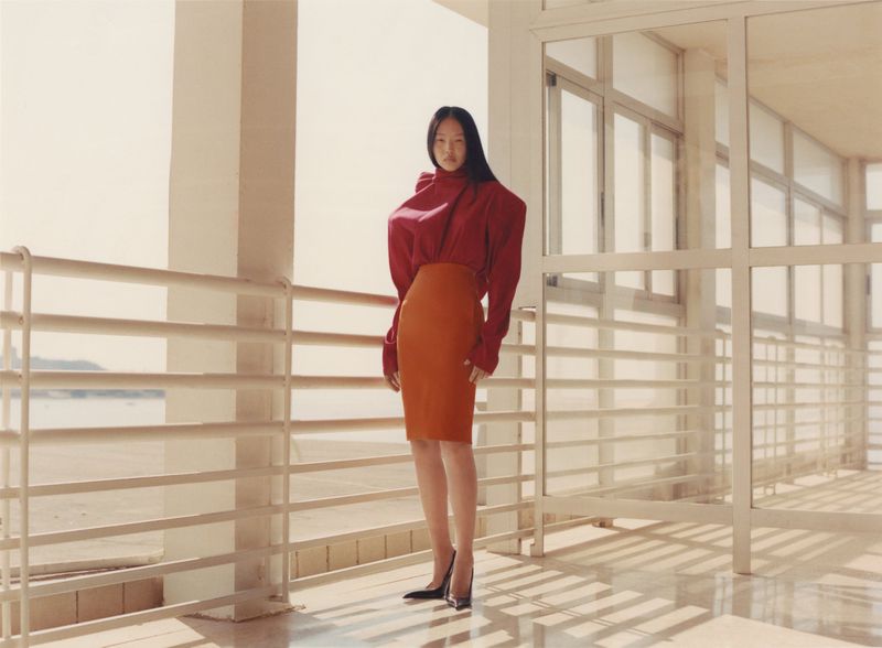 Fashion model in a red blouse and orange skirt poses in a bright indoor space with large windows and a sea view in the background.