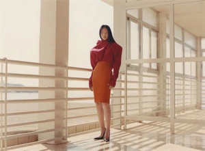Model Peng Chang poses on a sunlit architectural terrace wearing a draped red top, orange pencil skirt, and black pointed-toe heels.