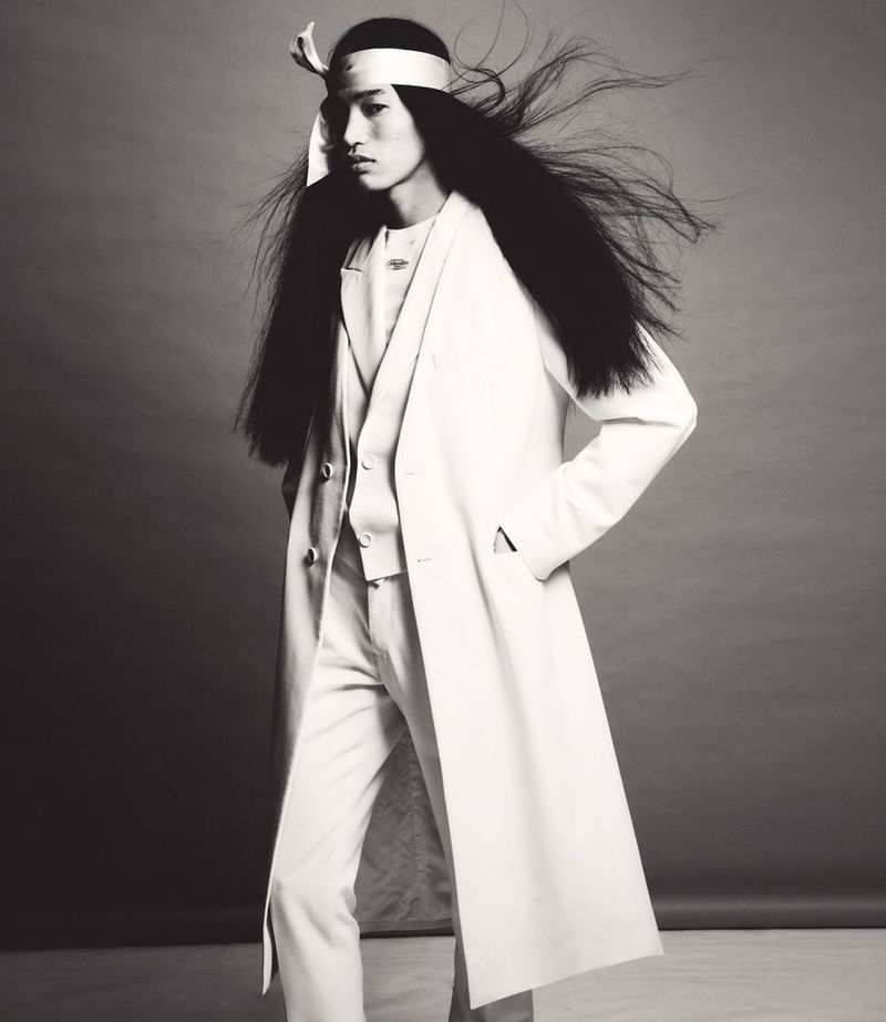 Fashion portrait of a young Asian male model with long hair and a white suit, standing in front of a neutral grey background.