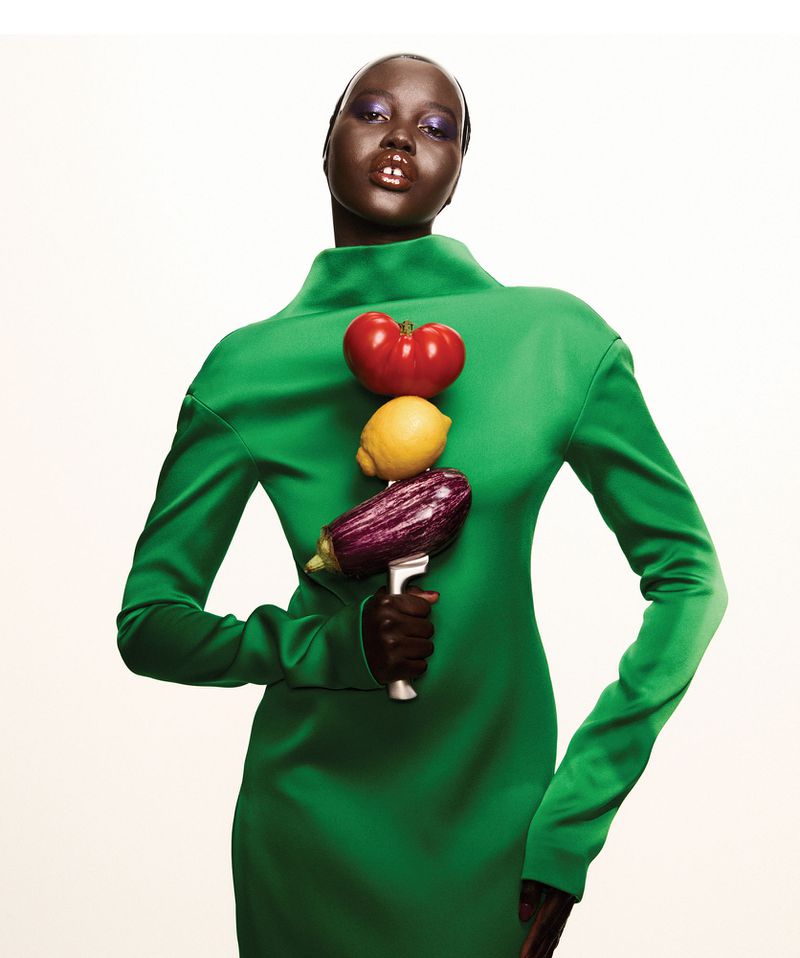 Fashionable woman in green dress poses with a stack of vegetables: tomato, lemon, and eggplant, against a clean, neutral background.