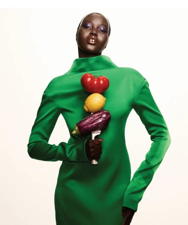 Fashionable woman in green dress poses with a stack of vegetables: tomato, lemon, and eggplant, against a clean, neutral background.
