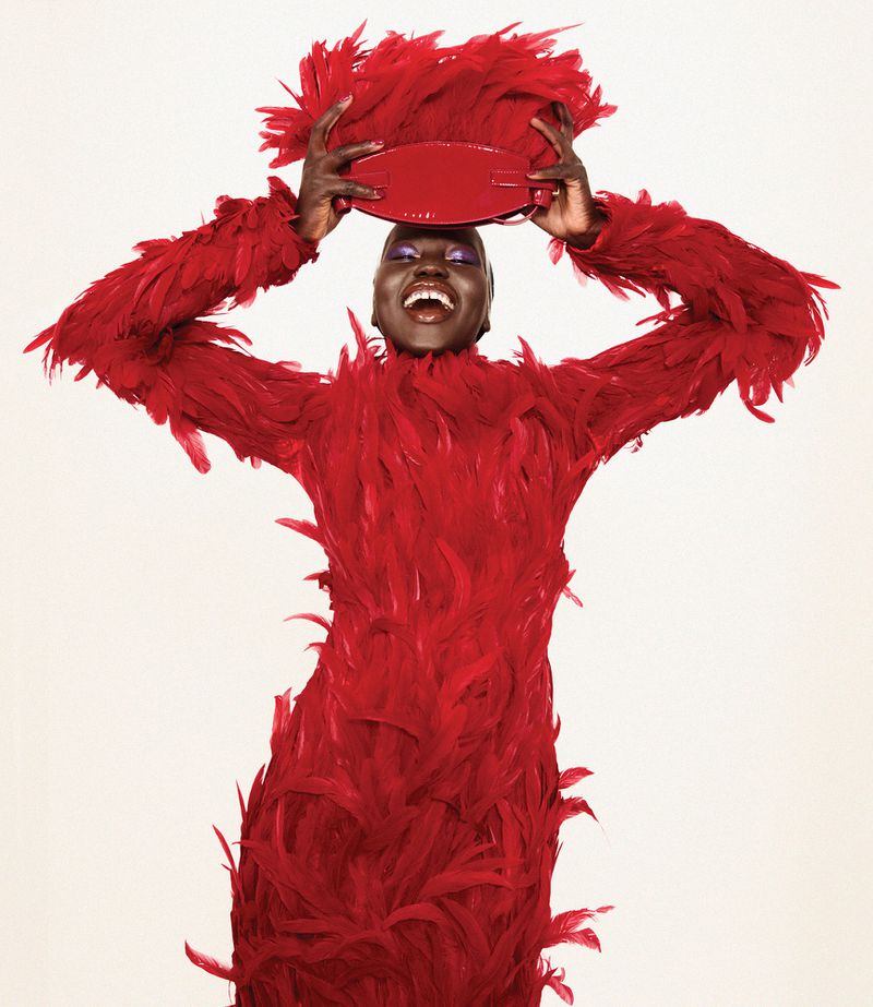 A smiling Black woman poses in a striking red feathered dress with a matching red purse held above her head against a white background.