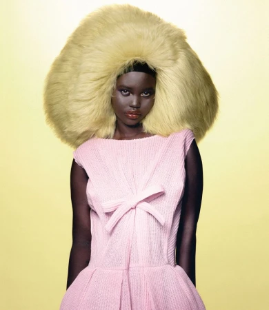 Dark-skinned woman with a large blonde wig and a pink dress posing against a yellow background in a fashion portrait, studio shot.
