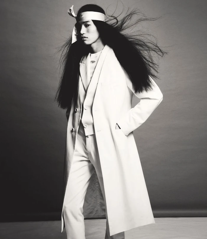Model with long dark hair wearing a white overcoat and headband against a neutral studio background.