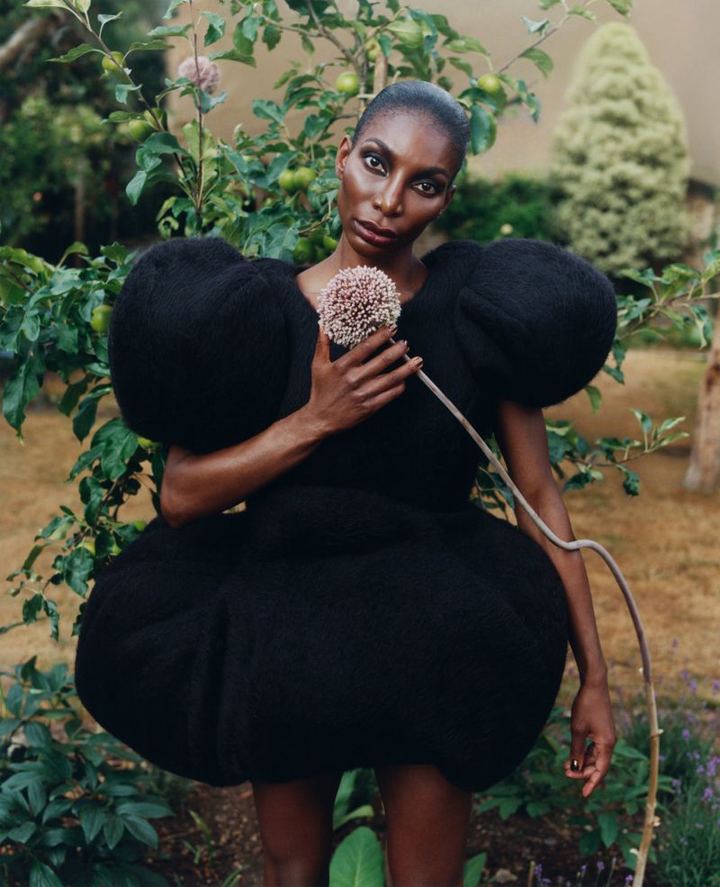 Elegant Black woman in a sculptural black dress holding a flower, standing in a garden. Fashion editorial shot with natural light.