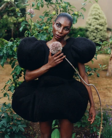 Elegant Black woman in a sculptural black dress holding a flower, standing in a garden. Fashion editorial shot with natural light.