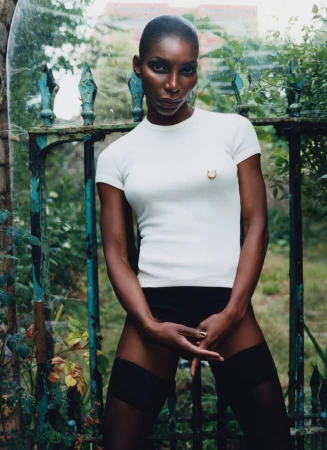 Elegant Black woman in a white top and black stockings stands confidently against a weathered garden gate and greenery backdrop.