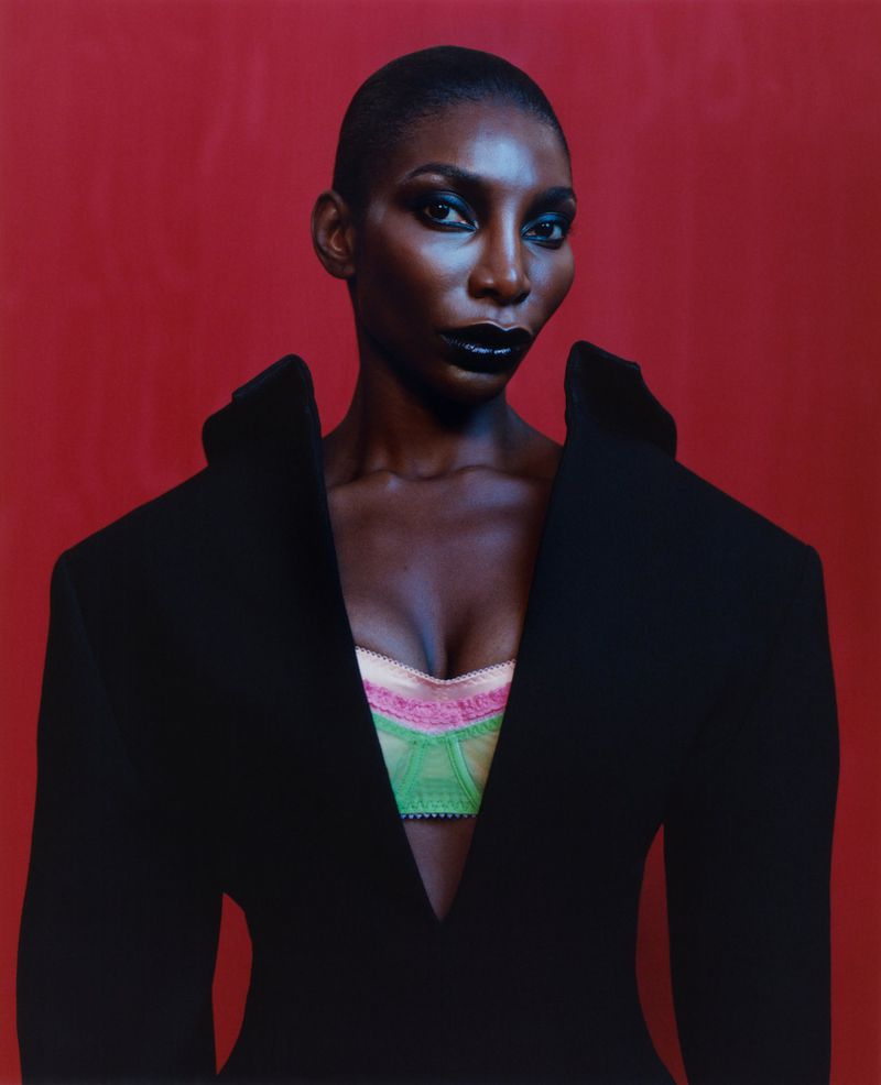 Close-up of a Black woman in a black jacket and colorful bra, set against a red background in a fashion-style portrait.