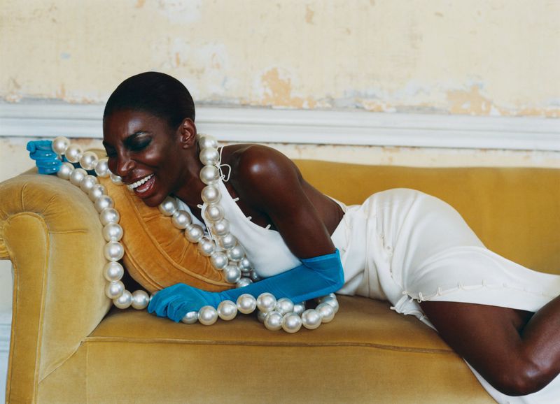 Woman in white dress lounges on yellow velvet couch with oversized pearl necklace and turquoise gloves, laughing in a bright indoor space.