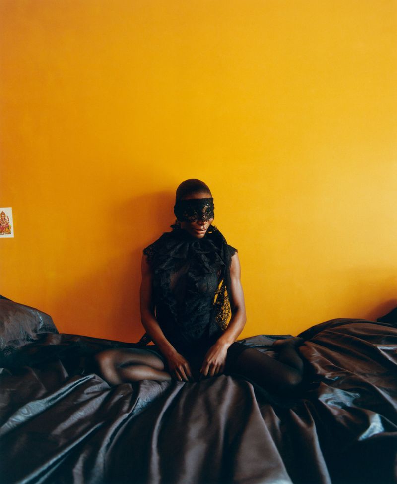 Woman in lingerie sitting cross-legged on a satin bed with a black lace blindfold in front of a bright yellow wall.