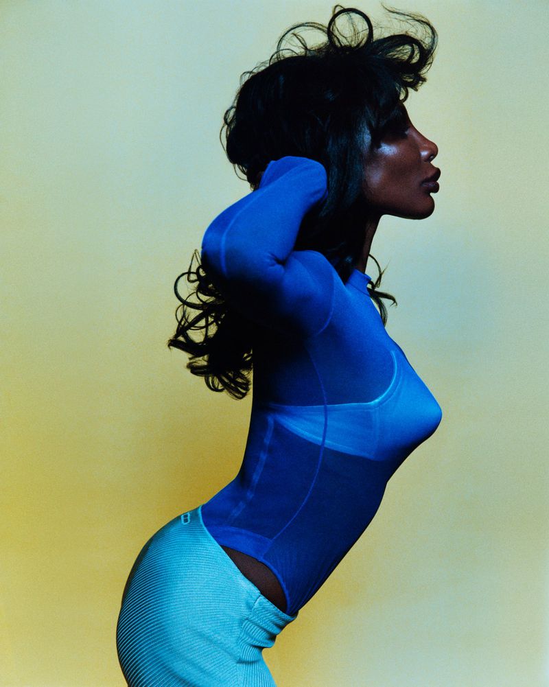 Portrait of an adult woman in a blue mesh top and blue skirt against a gradient yellow background in a studio setting.