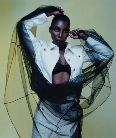 Fashion shot of a Black woman in a jacket, bra and skirt with sheer black fabric, posing in a photo studio against a muted yellow backdrop.