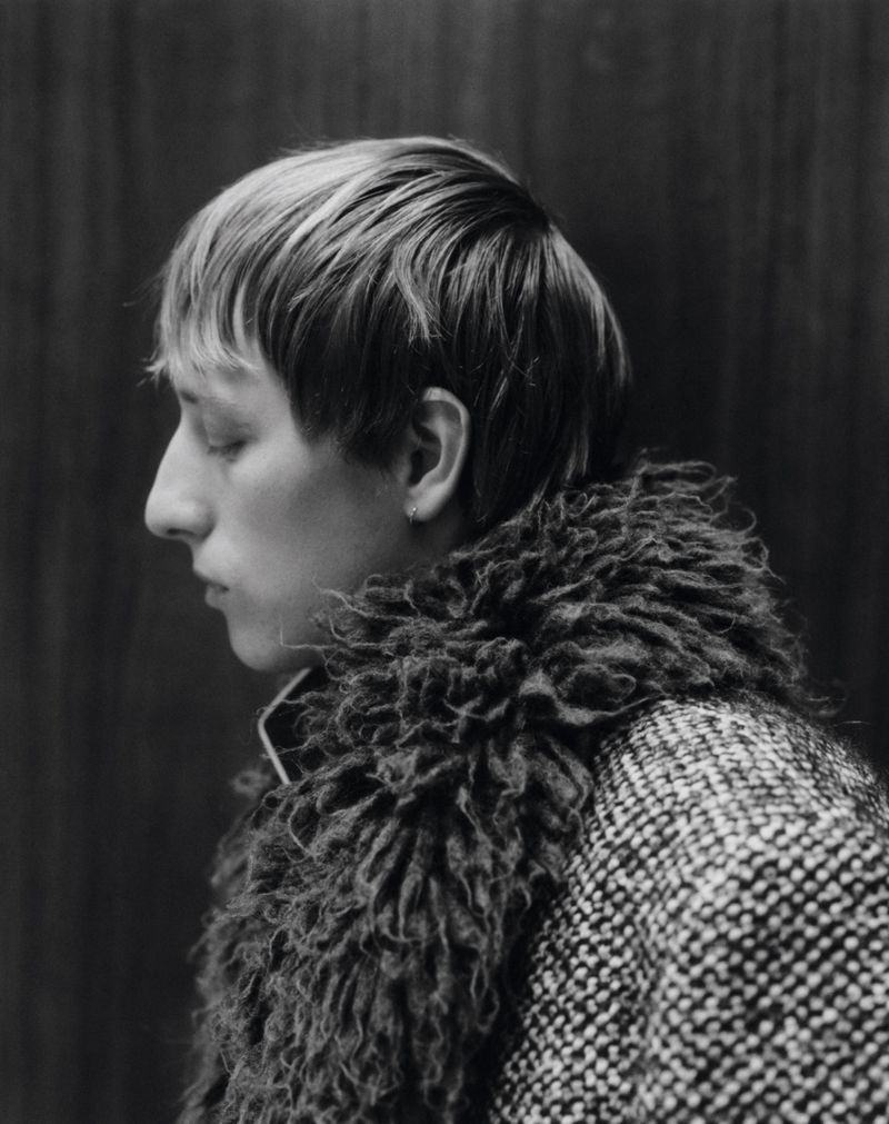 Black and white portrait of a young adult with eyes closed wearing a textured coat with a dark fur collar against a wood background.