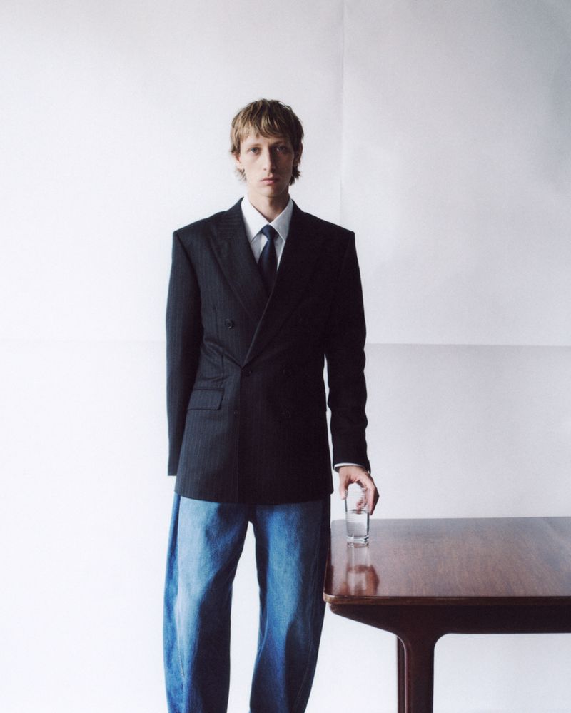 Portrait of a blond man in a dark business suit and jeans standing near a wooden table holding a glass in a white room.
