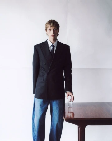 Portrait of a blond man in a dark business suit and jeans standing near a wooden table holding a glass in a white room.