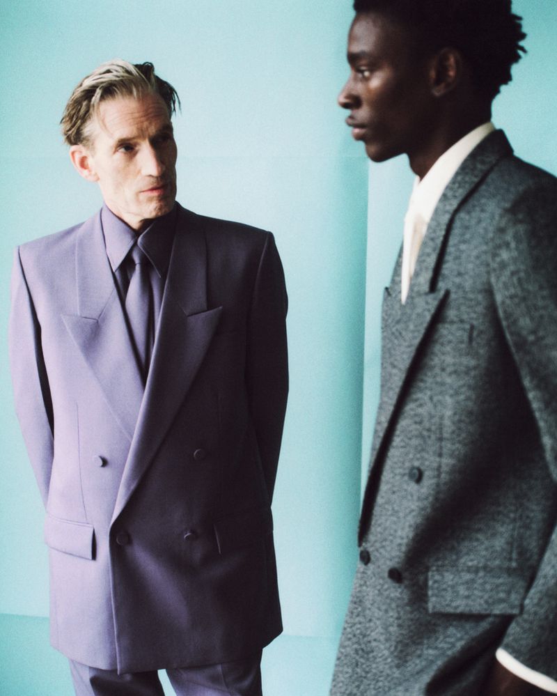 Two men in suits pose against a teal background. One wears a lavender suit and the other a grey suit in a fashion portrait.