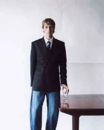 Model in black blazer and blue jeans standing beside a dark wood table holding a glass of water against a white background