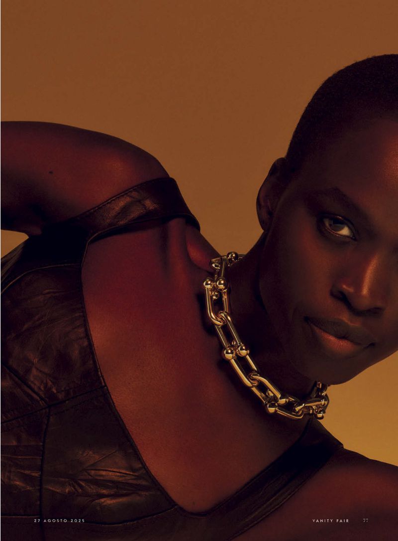 Close-up fashion shot of a dark-skinned woman with short hair wearing a chain necklace and dark top in a photo studio setting.