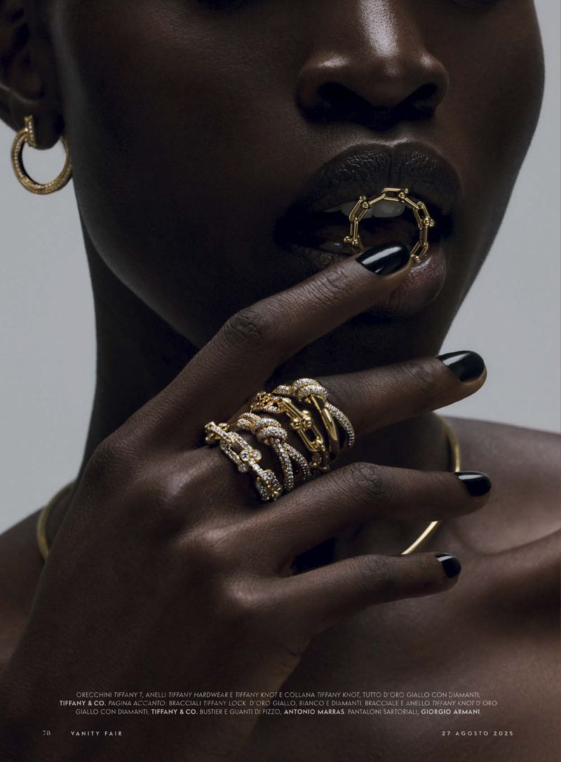 Fashion shot featuring a Black female model with glossy lips, wearing multiple rings and earrings in a studio setting with gold jewelry.