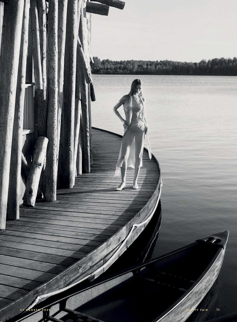 Monochrome image of a slender woman in a long dress standing on a wooden deck near a lake, with a canoe visible in the water.
