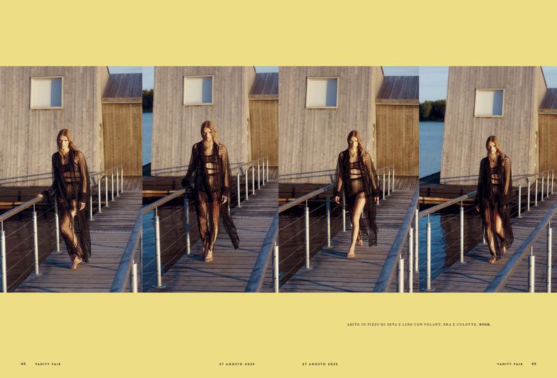 A woman walks on a wooden dock in a semi-sheer black dress and swimsuit, featuring a building and water in the background, editorial fashion.