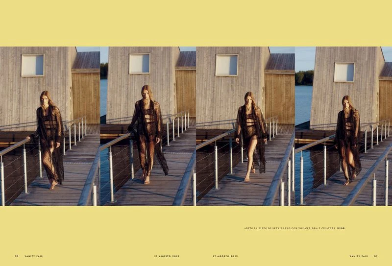A woman walks on a wooden dock in a semi-sheer black dress and swimsuit, featuring a building and water in the background, editorial fashion.