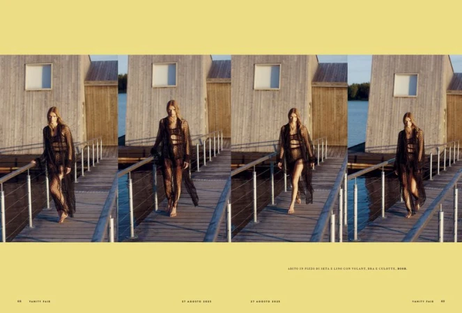 A woman walks on a wooden dock in a semi-sheer black dress and swimsuit, featuring a building and water in the background, editorial fashion.