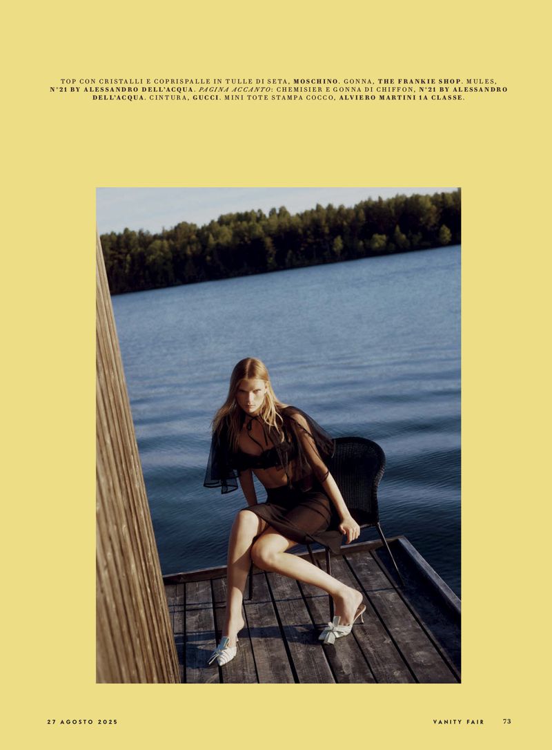 A blonde fashion model poses in sheer black clothing on a wooden dock, with a lake and trees visible in the background on a sunny day.