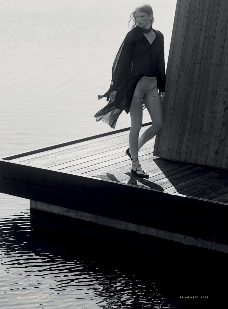 A woman poses on a wooden dock, wearing a sheer black dress and sandals, against a backdrop of water and a wooden structure.
