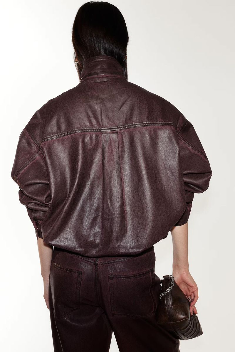 Back view of a woman wearing a plum-colored leather jacket and pants, with a matching handbag on a white backdrop in a fashion setting.