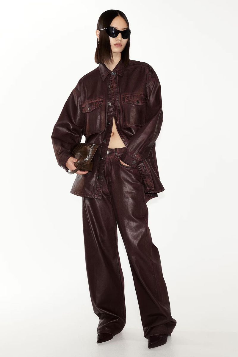 An Asian woman poses in a brown leather-like jacket and pants, holding a small bag, against a white studio backdrop for a fashion shoot.