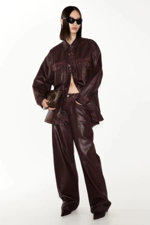 An Asian woman poses in a brown leather-like jacket and pants, holding a small bag, against a white studio backdrop for a fashion shoot.