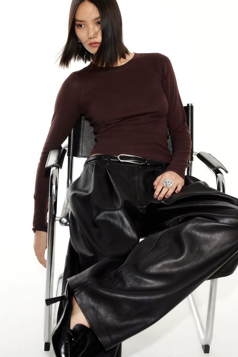 Model Huijia Chen seated in a modern chair wearing a dark brown long-sleeve top and black leather trousers against a white background