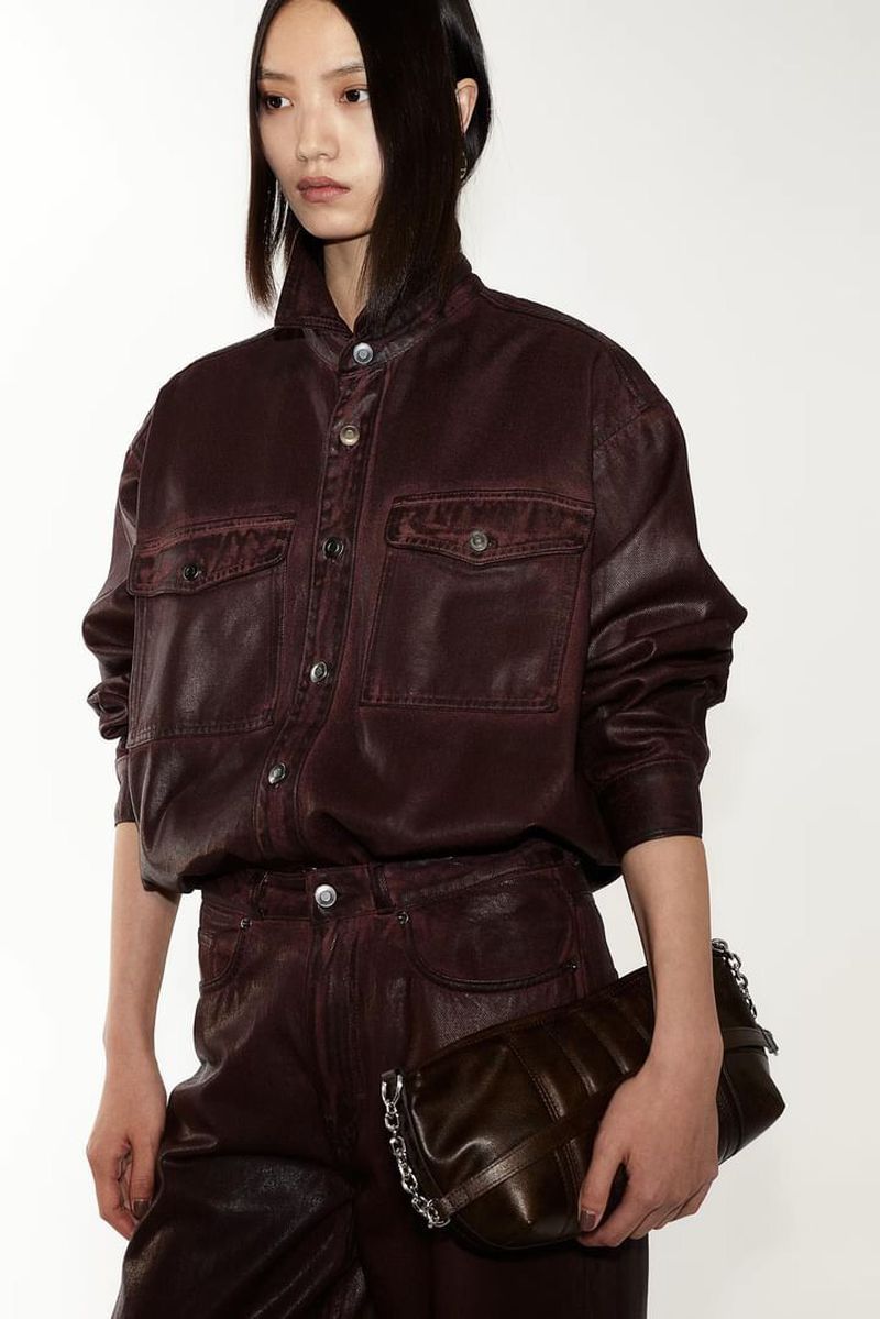 A fashionable Asian female model in a maroon leather-look shirt and pants, posing in a photo studio with a dark handbag.