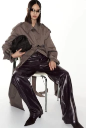 Model wearing dark sunglasses, grey coat, and glossy trousers seated on a chair against a plain background