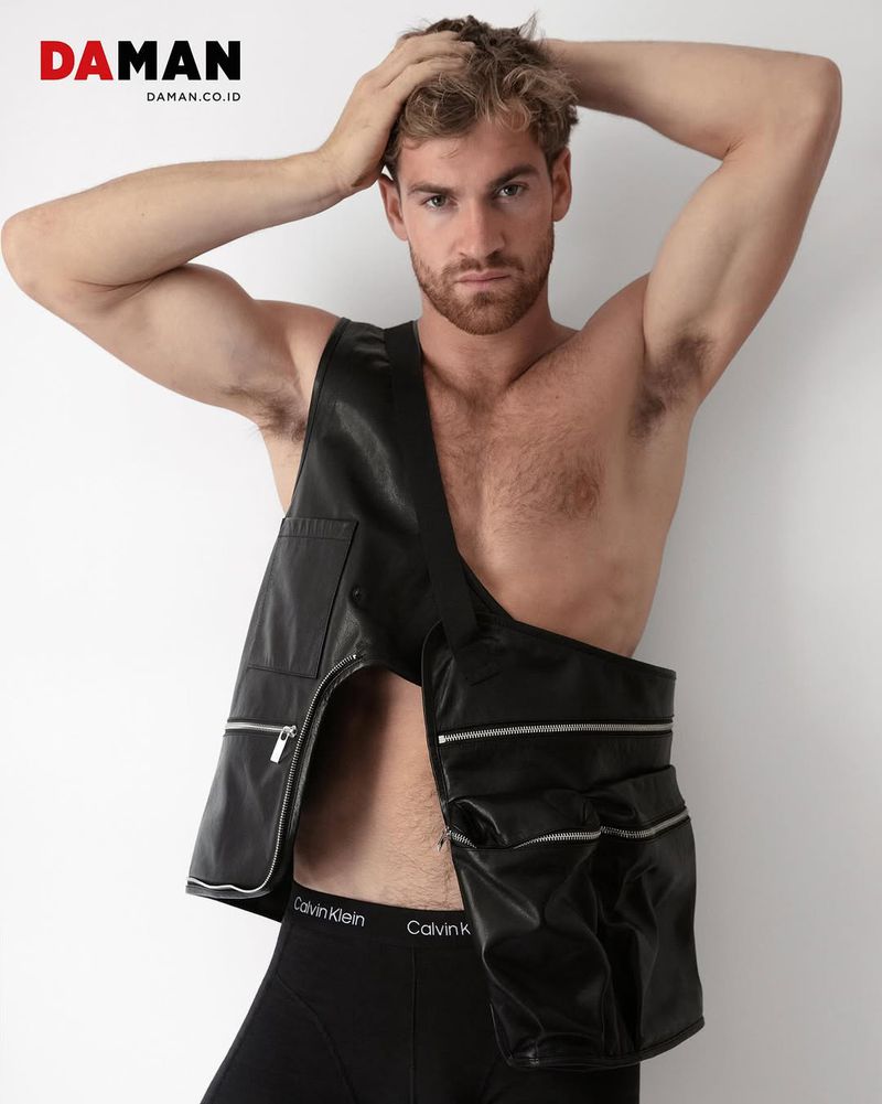 Male fashion model poses shirtless, wearing a black leather vest and briefs against a white backdrop in a studio shot.
