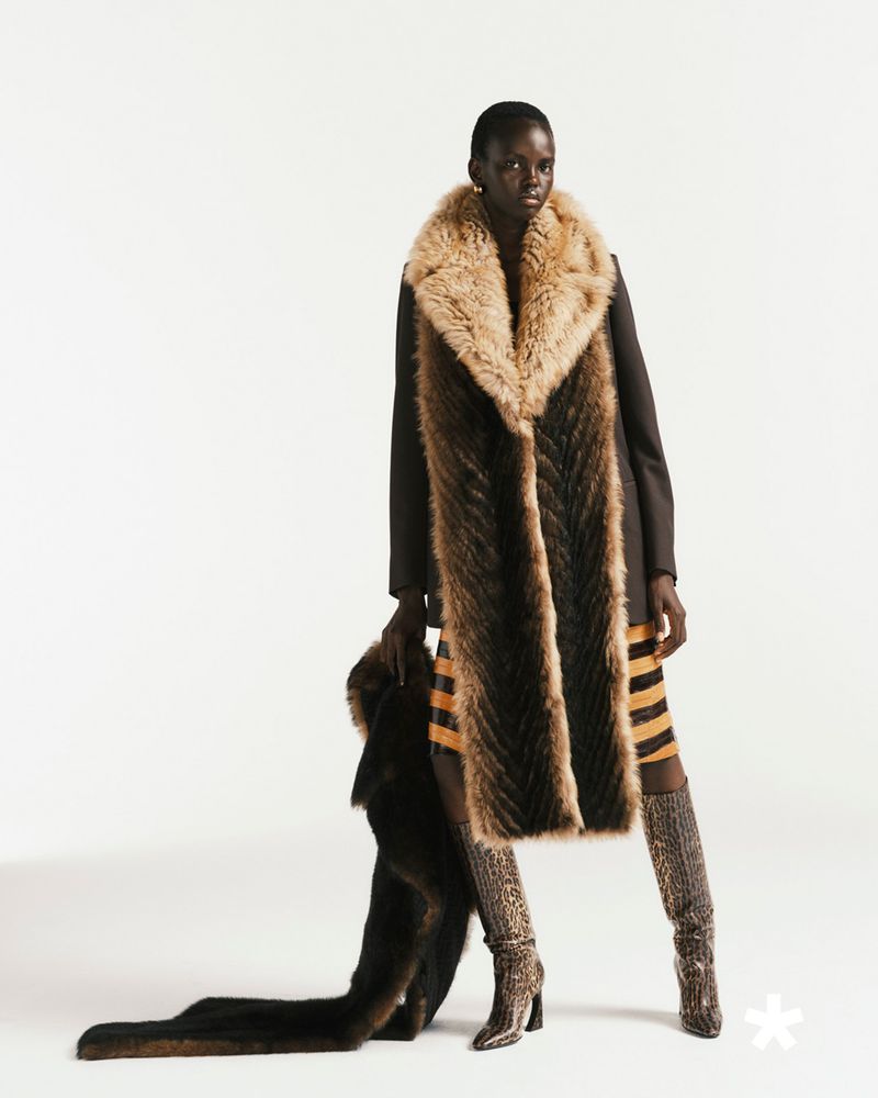 Fashion shot of a Black woman in brown coat and fur stole, posing in patterned boots against a white backdrop, showcasing fashion and style.