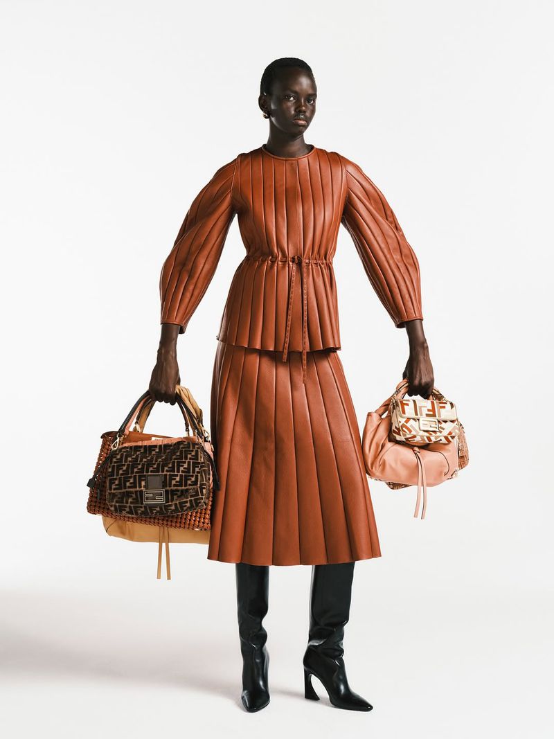 Fashion shot of a Black female model wearing a brown leather skirt and top while holding two Fendi bags on a white backdrop.