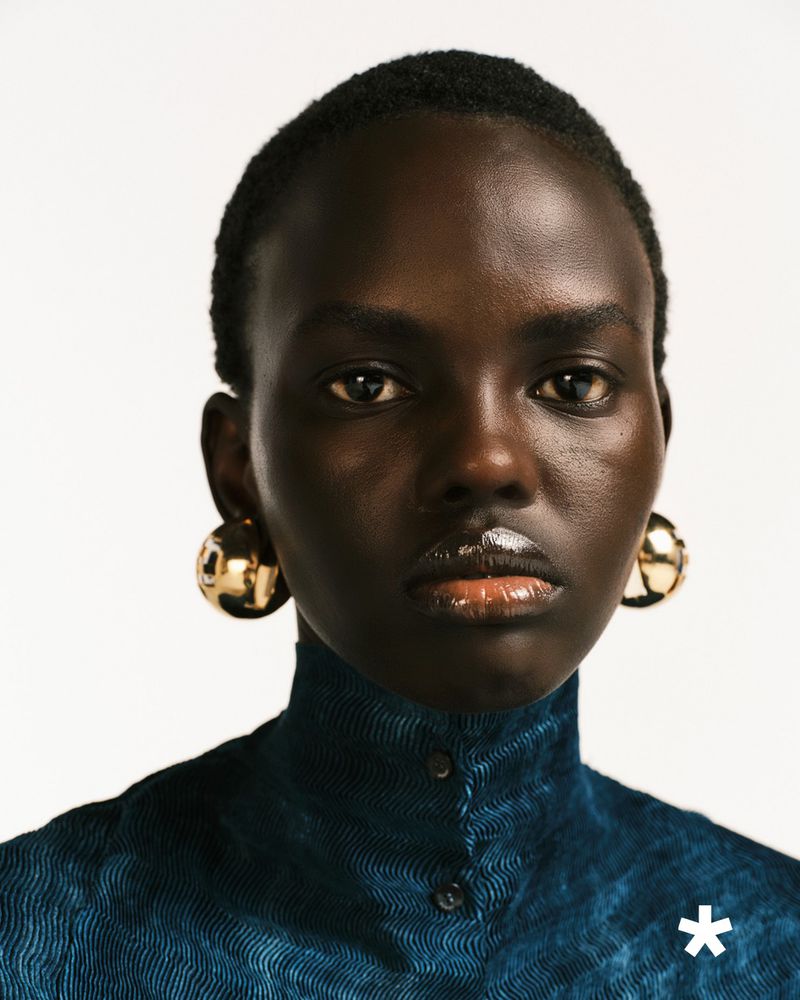 Close-up portrait of a striking dark-skinned woman with bold gold earrings, wearing a textured high-necked teal top against a white backdrop.