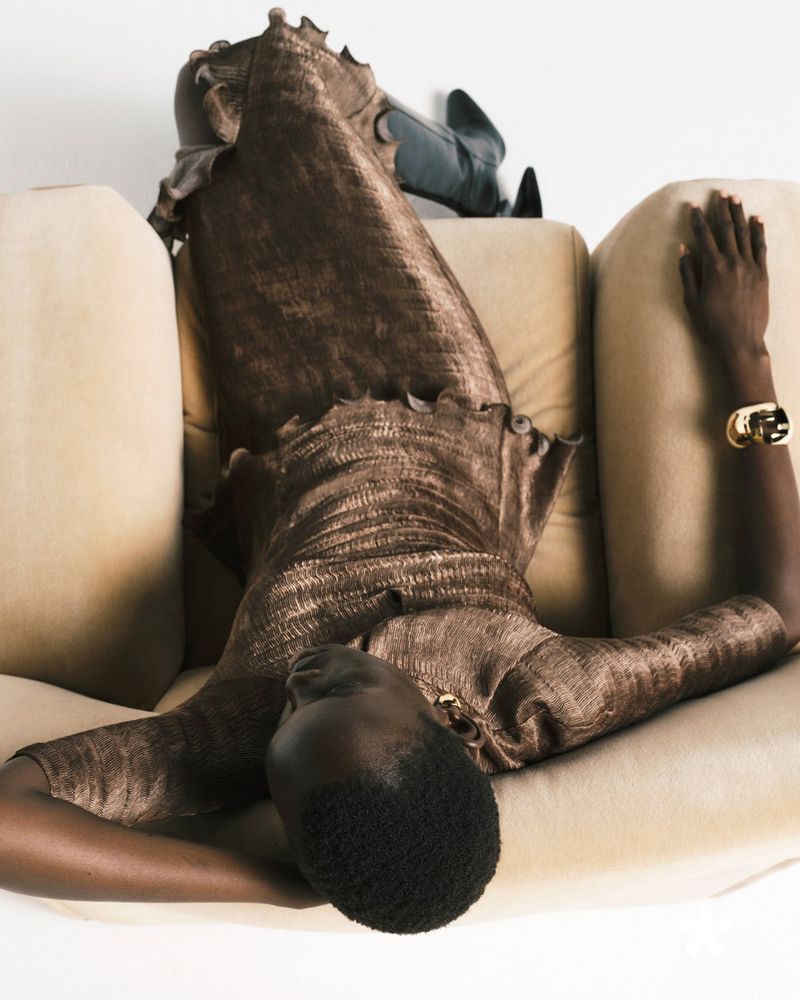 Dark-skinned female fashion model in brown textured ruffled dress rests on beige couch in studio. Bracelet visible. Fashion editorial.