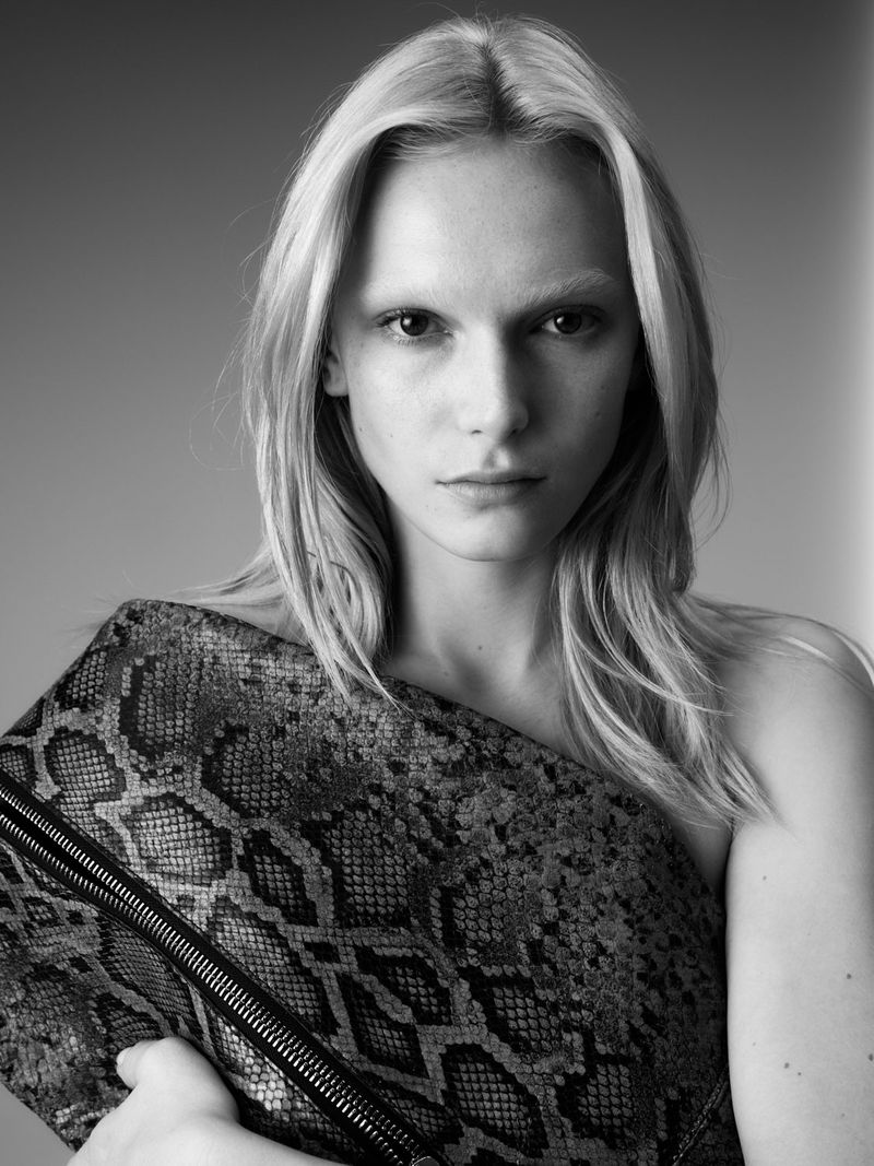 Monochrome portrait of a fair-skinned woman with blonde hair, holding a snakeskin textured clutch bag against a grey backdrop in a studio.