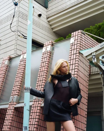 Fashion shot of a blonde woman in a black textured jacket and skirt, posing outdoors against a building with security cameras.