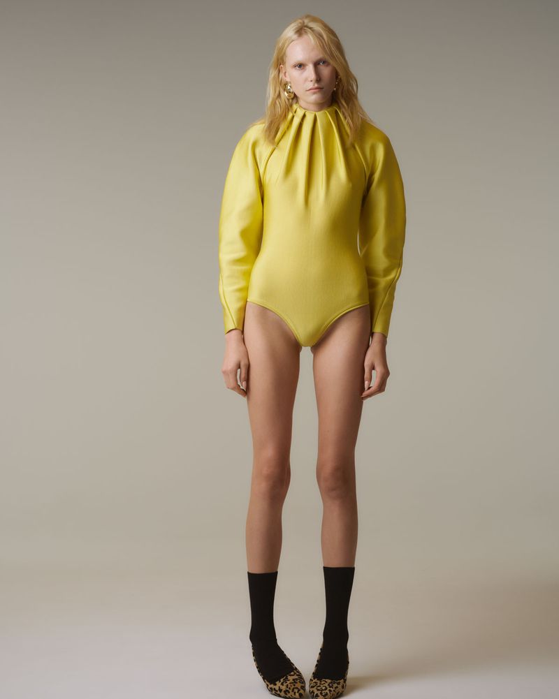 Studio shot of a slender fashion model in a yellow bodysuit with puffy sleeves, black socks, and leopard print heels. Full body.